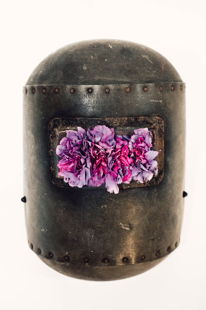 photograph of welder's mask filled with purple flowers where eyes should be